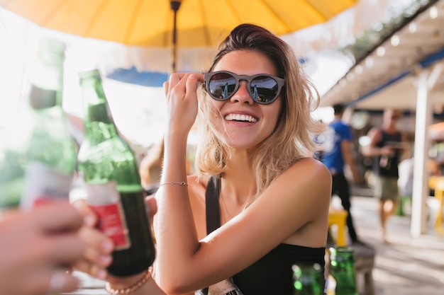 Смеющаяся беззаботная девушка пьет пиво в летнем кафе. Красивая загорелая дама в солнечных очках с удовольствием позирует в теплый день.