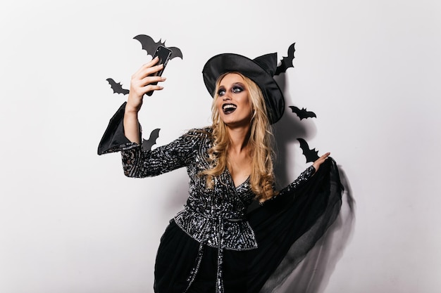 Смеющаяся беззаботная девушка делает селфи с хэллоуинскими летучими мышами Студийный портрет приятной молодой ведьмы, танцующей на вечеринке