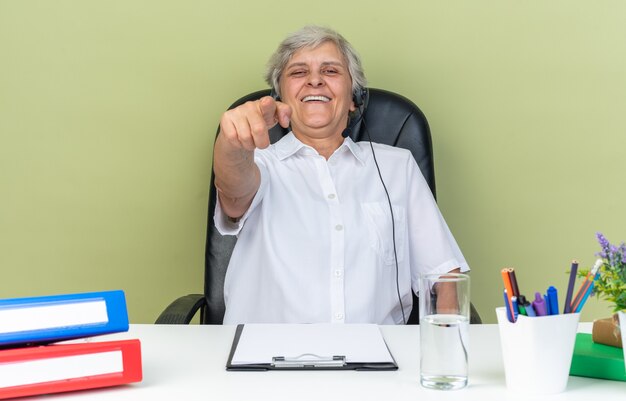 녹색 벽에 격리된 사무실 도구를 들고 책상에 앉아 있는 헤드폰을 끼고 웃고 있는 백인 여성 콜센터 교환원