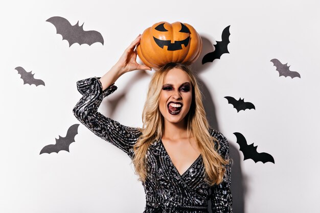 Laughing cute vampire with blonde hair holding pumpkin. Gorgeous white woman in witch attire having fun in halloween.