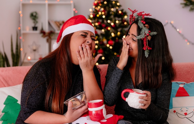 laughing daughter and mother covering mouth with hand sitting on couch enjoying christmas time at home