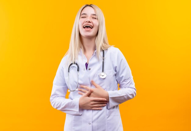 Laughing doctor young girl wearing stethoscope in medical gown and dental braces put her hands on stomach on isolated yellow wall