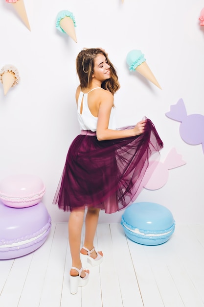 Laughing dreamy girl in white tank-top playing with her lush skirt, standing near the colorful toy macaroons. Pretty elegant young woman in romantic outfit dancing on wedding party.