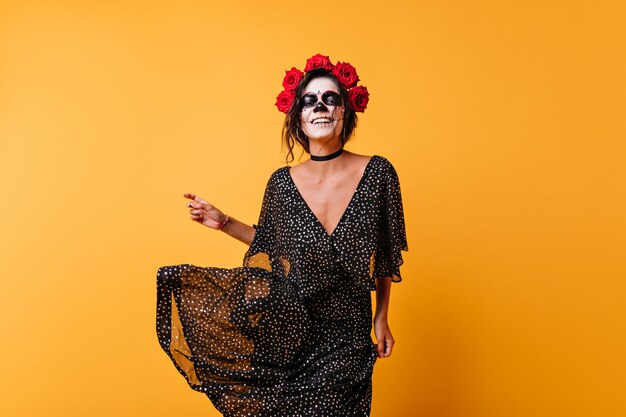 Free photo laughing female zombie with roses in hair dancing in studio. happy girl with mexican makeup celebrating halloween.