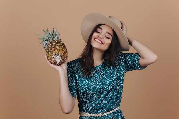 La ragazza ridente in cappello e vestito verde posa con un ananas nello studio