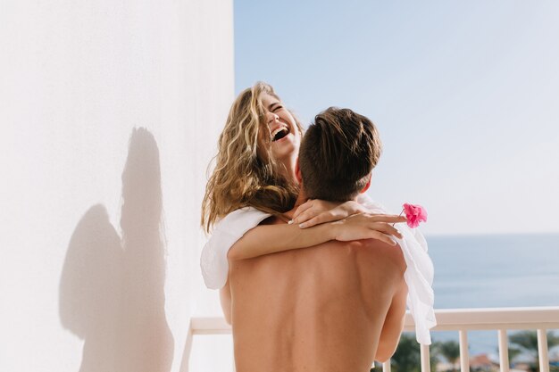 그녀의 남자 친구와 함께 여름 휴가를 보내는 테라스에서 재미 흰 블라우스에 웃는 소녀. 이국적인 리조트에서 신혼 여행에 바다 전망 발코니에서 포옹하는 아름다운 행복한 커플