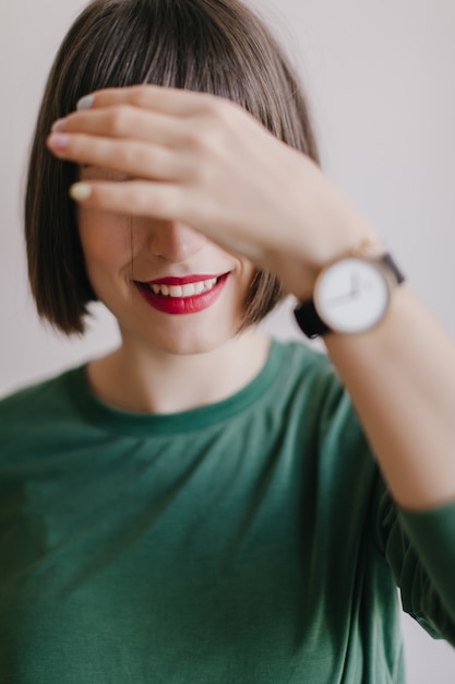 赤い唇で笑っている女の子は彼女の目を覆います。トレンディな腕時計のポーズで快適な女性モデルの屋内写真。