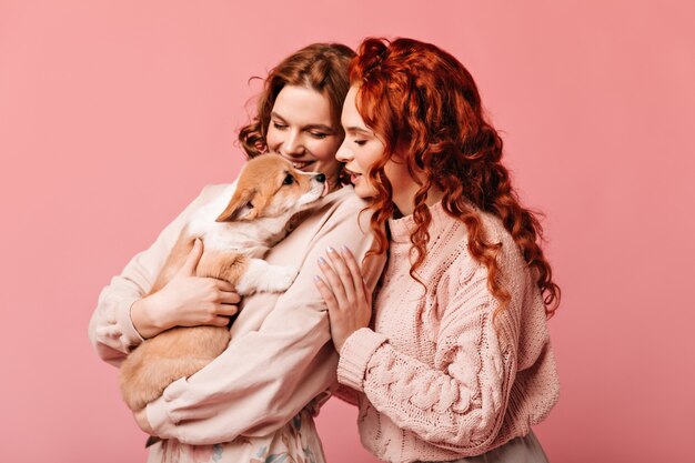 Laughing girls looking at puppy. Studio shot of adorable ladies with dog posing on pink background.