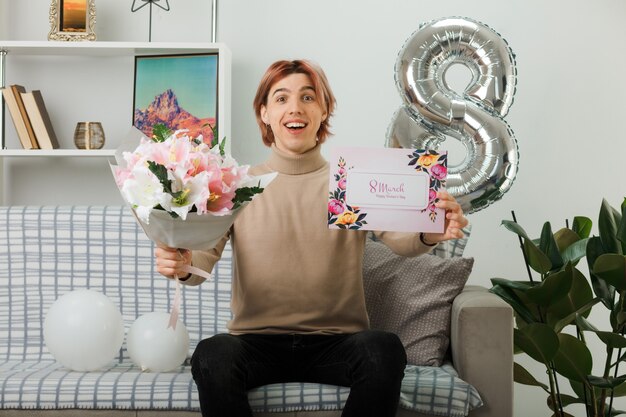 Laughing handsome guy on happy women day holding bouquet with greeting card sitting on sofa in living room