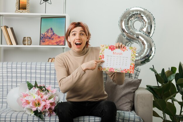 Laughing handsome guy on happy women day holding and points at calendar sitting on sofa in living room