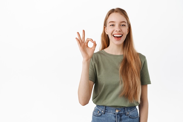 Laughing happy candid girl, showing okay OK sign and nod in approval, agree, like and praise something good, make compliment, recommend excellent product, standing over white wall