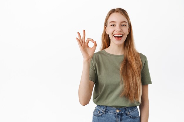 Laughing happy candid girl, showing okay OK sign and nod in approval, agree, like and praise something good, make compliment, recommend excellent product, standing over white wall