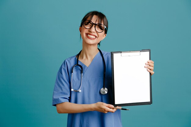 青い背景で隔離の均一なフィス聴診器を身に着けているクリップボードを持って笑う若い女性医師