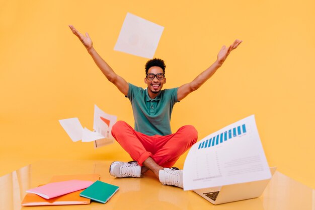 Laughing international student throwing out papers. Indoor photo of african male freelancer sitting on the floor with laptop.