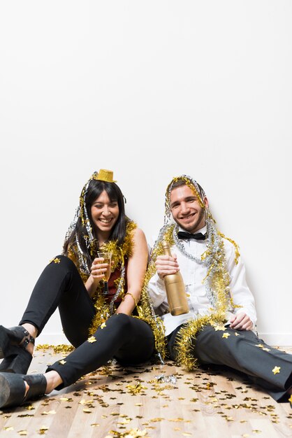 Laughing lady and guy in evening wear and tinsel with bottle and glass on floor 