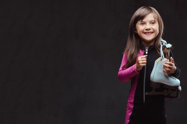 Laughing little girl dressed in sportswear holds ice skates on a shoulder. Isolated on dark textured background.