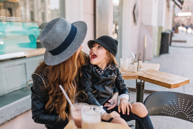 Смеющаяся маленькая девочка в черной шляпе и куртке стоит на коленях мамы и дурачится.