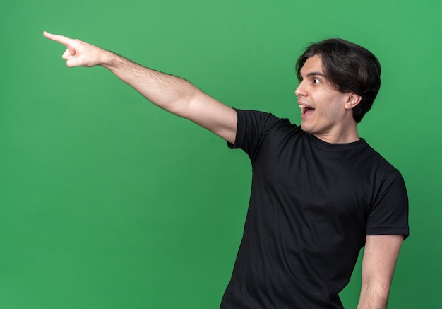 Laughing looking at side young handsome guy wearing black t-shirt points at side isolated on green wall