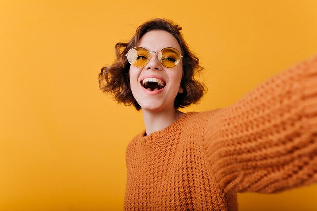 Selfie를 만드는 재미있는 노란색 선글라스에 멋진 여자를 웃 고있다. 자신의 사진을 찍는 니트 스웨터에 편안한 백인 여자의 초상화.