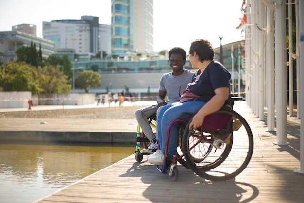 Free photo laughing man and woman in wheelchairs spending time near water. african american man and caucasian woman in casual clothes, looking at water, joking. love, affection, happiness concept