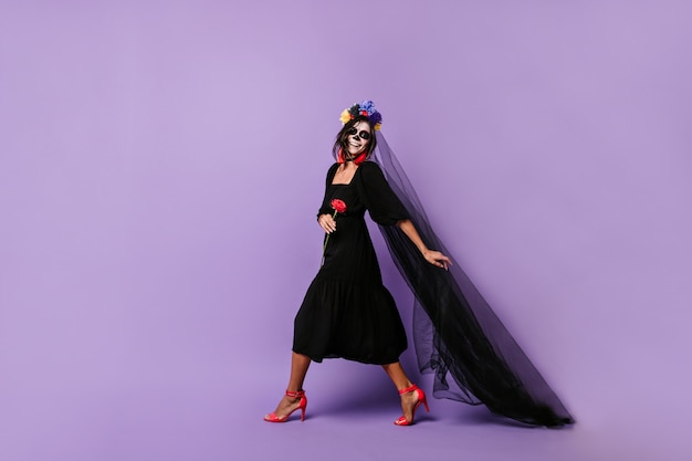 Бесплатное фото Смеющаяся мексиканская модель в костюме хэллоуина идет через сиреневую стену, держа в руках длинную черную вуаль.