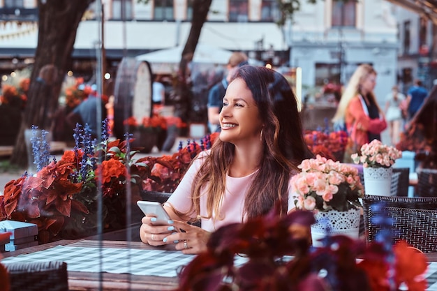 Laughing middle age businesswoman with long brown hair holds a smartphone while sitting at an outdoor cafe.