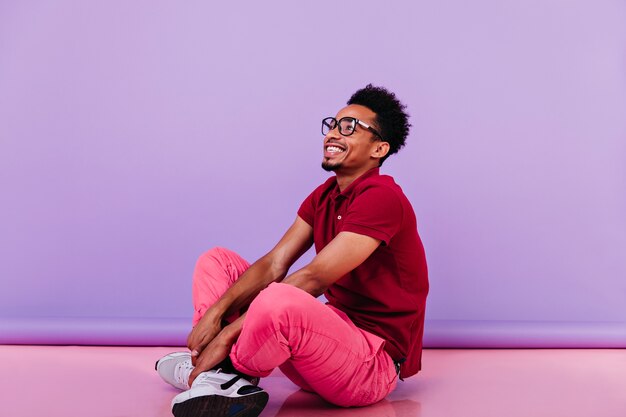 ピンクのズボンに座って楽観的な男を笑う。幸せな笑顔で床にポーズをとる感情的な黒人の若い男。