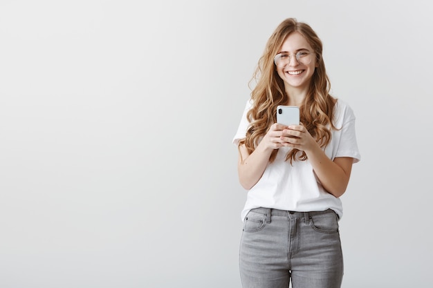 撮った写真の女の子を笑って。メガネとスマートフォンを保持しているトレンディな服装で幸せな魅力的な白人女性ブロガーの肖像画、肯定的な感情から笑う