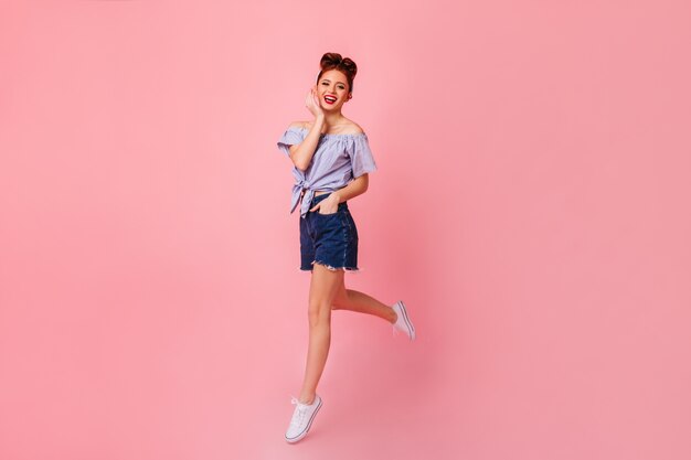 Laughing pinup girl posing with hand in pocket. Full length view of pretty ginger woman in denim shorts jumping on pink space.