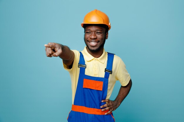 Laughing points at side young african american builder in uniform isolated on blue background