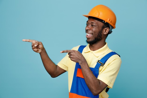 Laughing points at side young african american builder in uniform isolated on blue background