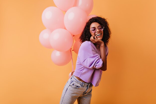 Laughing pretty black woman posing with party balloons. Inspired birthday girl with curly hair standing on orange.