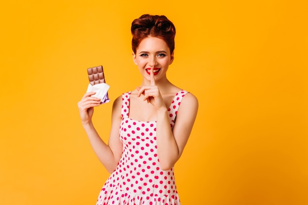 초콜릿을 들고 폴카 도트 드레스에 예쁜 아가씨를 웃고 있습니다. 노란색 공간에 포즈 행복 핀 업 소녀의 스튜디오 샷.
