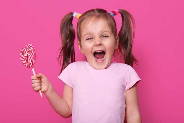 Laughing screaming cute girl opens her mouth widely, showing teeth, holds in one hand heart bright tasty lollipop. Emotional positive childen spends her leisure time with pleasure. Copy space for ad.