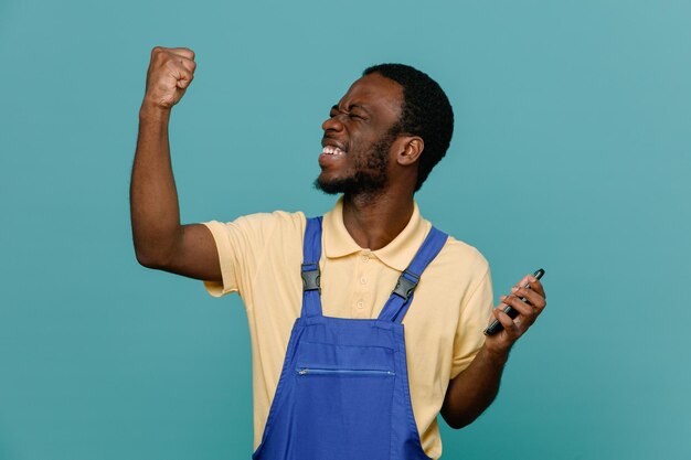 Смеясь, показывая сильный жест, держа телефон молодой афроамериканец уборщик мужчина в униформе, изолированные на синем фоне