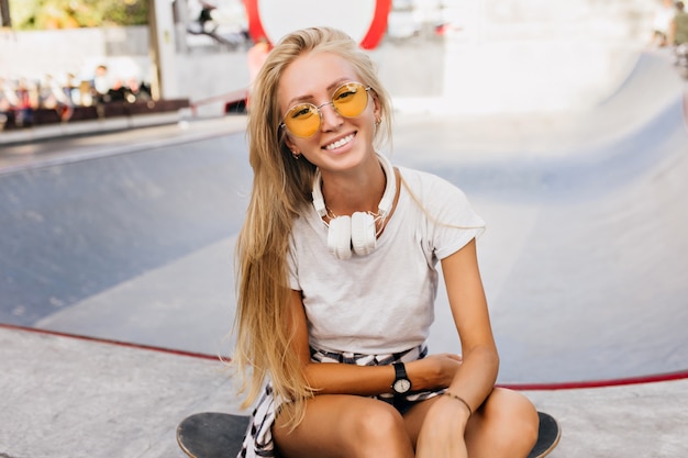 Donna pattinatrice di risata in occhiali da sole gialli in posa all'aperto. foto di signora dai capelli biondi estatica che si siede sullo skateboard nel parco.