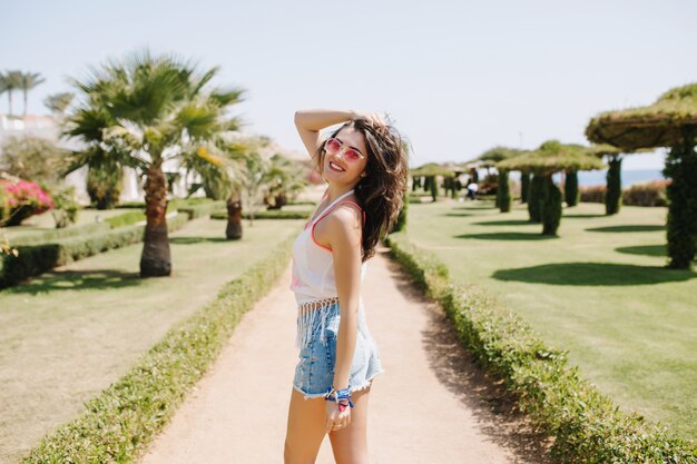 Laughing slim girl in trendy sunglasses with dark hair spending time outside, walking in the park in summer morning. Amazing young woman in denim shorts smiling, while posing on palm tree