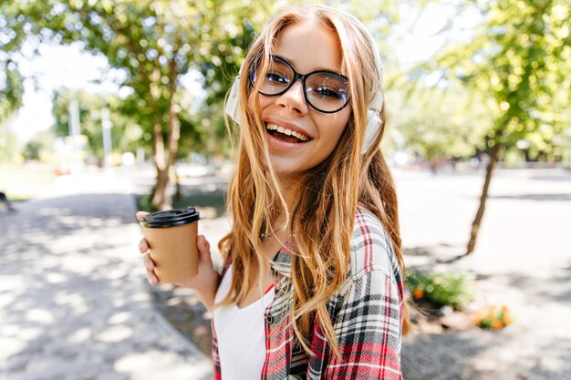 公園でお茶を楽しんで笑う壮大な女性。自然の中でコーヒーを飲むスタイリッシュな白人の女の子。