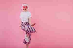 Бесплатное фото Смеющаяся потрясающая женщина в солнечных очках танцует на розовой стене. веселая европейская модель в гламурном перуке развлекается