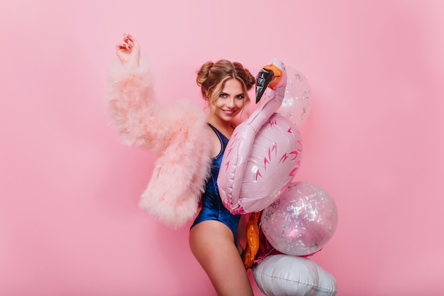 Laughing stylish girl in fluffy pink jacket dancing with event balloons, waiting for friends. Adorable young woman in blue bodysuit having fun with toy flamingo, ready for pool party.