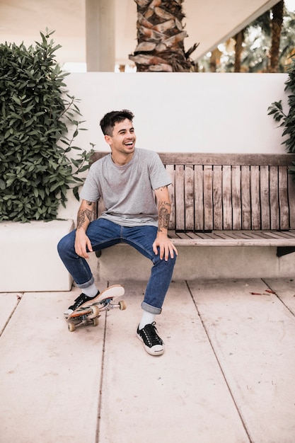 Laughing teenage boy sitting on wooden bench by placing foot on skateboard