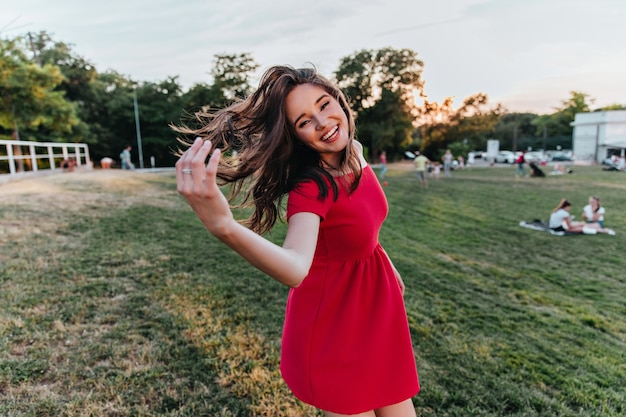 Laughing winsome woman with dark hair posing . Carefree female model in red dress dancing in park.