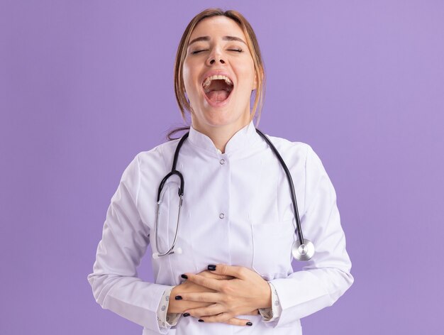 Laughing with closed eyes young female doctor wearing medical robe with stethoscope grabbed stomach isolated on purple wall