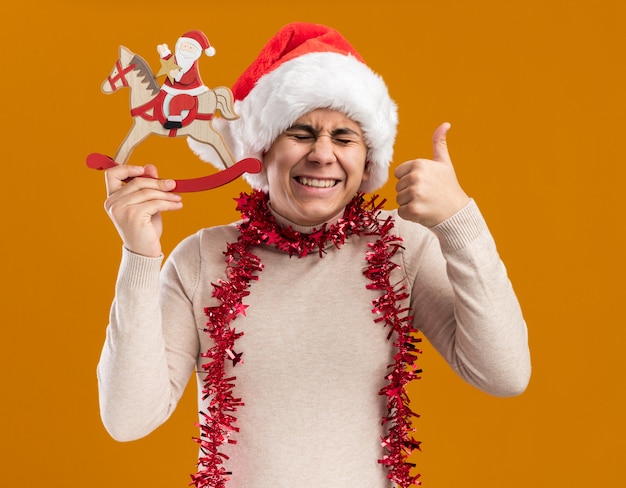 Laughing with closed eyes young guy wearing christmas hat with garland on neck holding christmas toy showing thumb up isolated on yellow wall