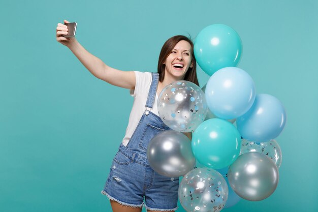 파란색 청록색 벽 배경에 격리된 다채로운 공기 풍선을 들고 축하하는 휴대폰으로 셀카 촬영을 하는 데님 옷을 입은 웃고 있는 여성. 생일 휴일 파티, 사람들의 감정 개념입니다.