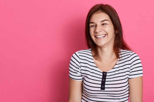 Laughing woman with dark hair posing isolated over rosy wall, happy girl wearing striped t shirt, expressing happiness and joy. Copy space for advertisement.