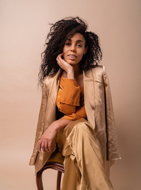 Laughing woman with perfect curly hair in elegant orange blouse and silk pants sitting on vintage chair