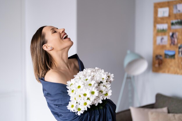 Laughing woman wrapped in a blue bedsheet holding a bouquet of white flowers in the living room Happy people