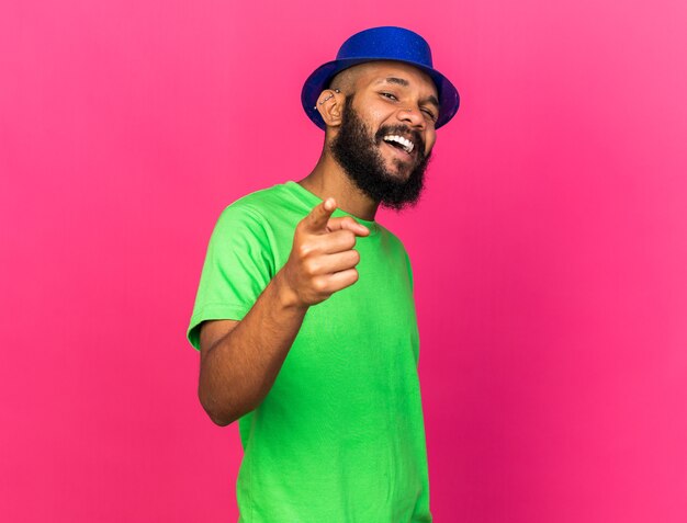 Laughing young afro-american guy wearing party hat points at front isolated on pink wall