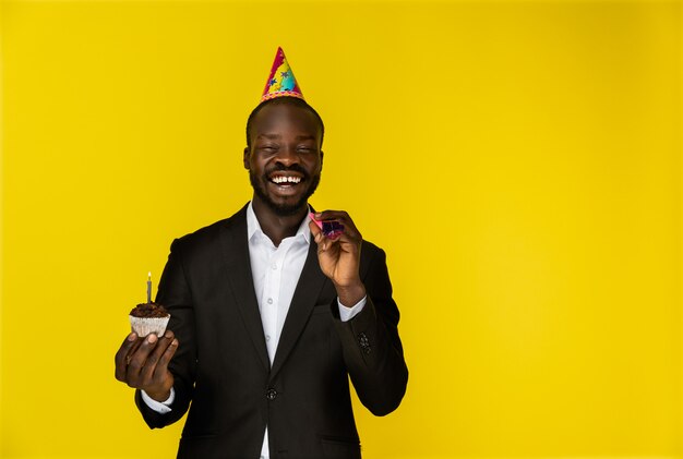 Laughing young afroamerican guy in black suit and birthday hat with burning candle 